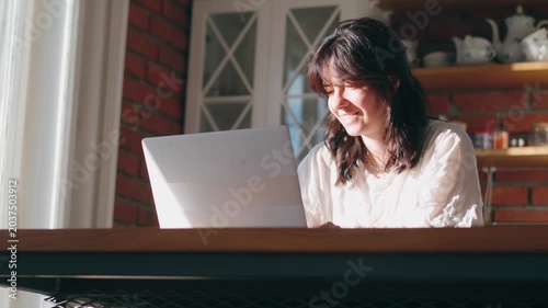 Portrait of a young woman working on a laptop at a kitchen table. Remote work, home office, focused professional, modern lifestyle, productivity in a cozy home interior.
