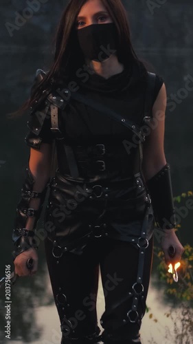  fantasy woman warrior assassin holds burning fire dagger in hands walks at night nature. Gothic sexy armed girl. Black leather costume mask hides face. Ninja sword knife. 4k footage. river rocks