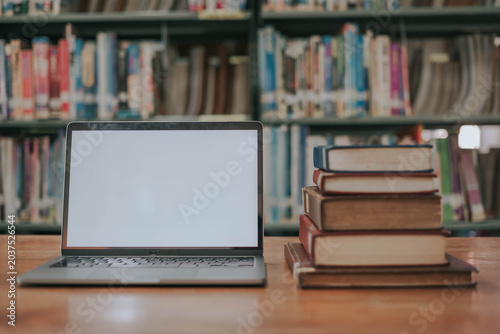 A simulated image of an empty laptop screen on a wooden table with a stack of books in a library, with a library as the backdrop.