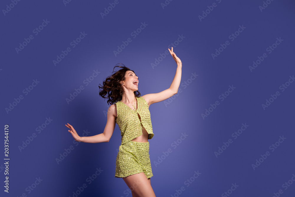 Naklejka premium Joyful young woman dancing and leaping in a green summer outfit against a purple studio background, expressing happiness, movement and carefree energy