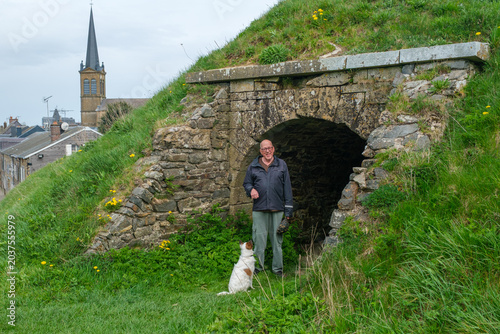 senior man stands by a bunker in village of Rocroi in France; his dog is sitting with him.