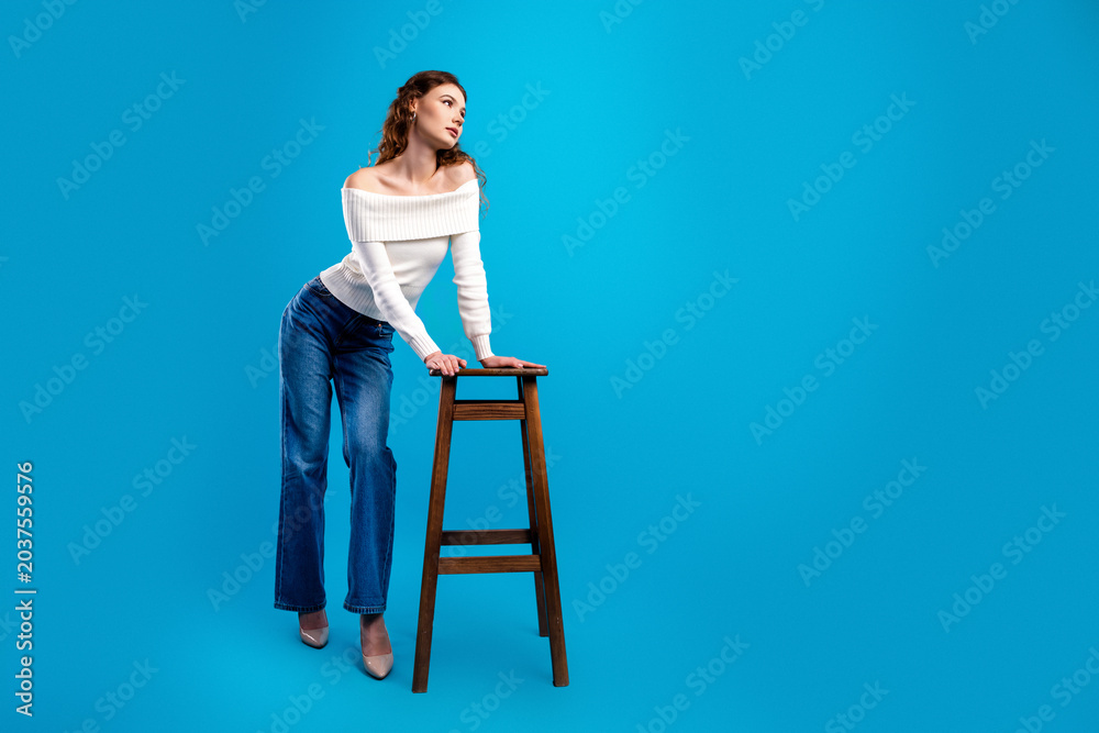 Fototapeta premium Fashionable woman in off shoulder white sweater and blue jeans posing elegantly with a wooden stool on a bright blue studio background