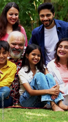Indian family sitting on lawns smiling at camera, three generations including grandparents parents and children bonding together outdoors in park, happy Asian family enjoying leisure time together