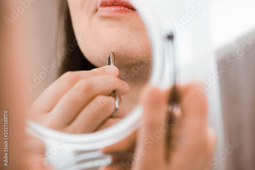 Photos Reflection in a mirror of a woman using tweezers for facial hair removal on her chin, managing unwanted hair and hormonal changes in her home beauty routine