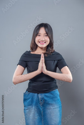 A smiling Asian woman with hands together in a greeting gesture, wearing a black top and denim skirt, expressing friendliness, respect, and confidence with a cheerful and welcoming attitude.
