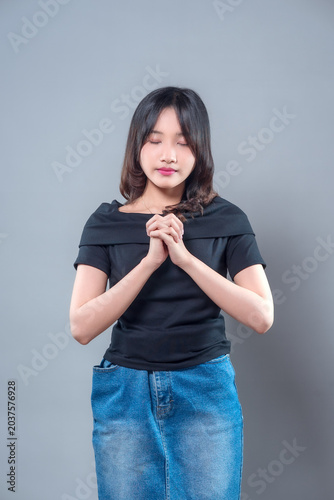 An Asian woman with closed eyes and a calm expression holding her hands together, wearing a black top and denim skirt, showing peace, mindfulness, and graceful beauty in a serene moment.