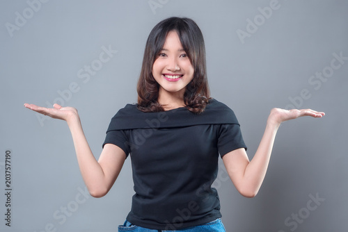 A cheerful Asian woman wearing a black top and denim skirt smiling with both hands raised, showing a welcoming and confident gesture, suitable for business, comparison, or advertising concepts.