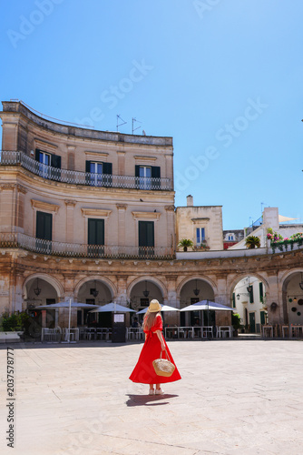 Woman in red dress walking through sunny European square in Martina Franca with historic architecture, summer travel atmosphere and vacation lifestyle