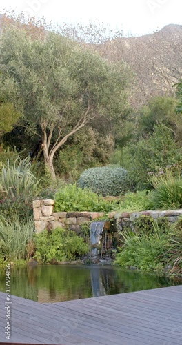 Vertical video: Gravity pulling cascade over stone lip in garden and stirring pond ripples