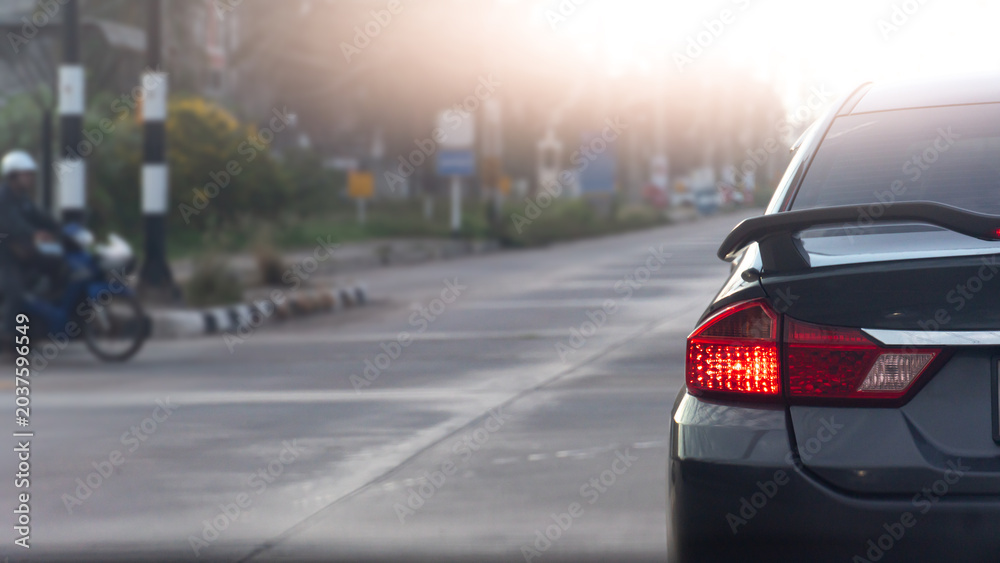 Fototapeta premium Rear corner close up of a dark car showing its illuminated red tail light. Car is stopped on a wide city road with soft daylight. Blurred of motorcycles waiting to cross the intersection.
