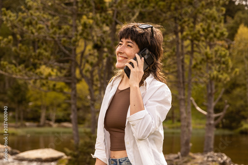 Woman talks on her phone in a sunlit park. She smiles, enjoying nature by a lake. Trees and rocks frame the peaceful setting. She wears a white jacket and jeans. Sunglasses rest on her head