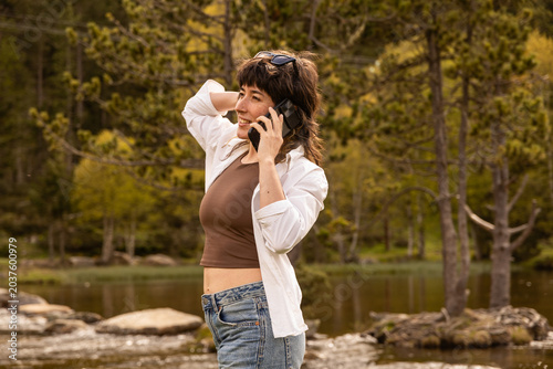 Woman stands near a river. She wears a white shirt and blue jeans. Her hair is tied up. She holds a phone to her ear. She looks relaxed and happy. This image works for travel or nature photography
