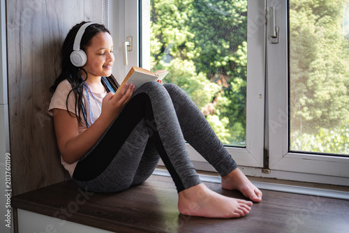 Young girl reading book and listening music by window