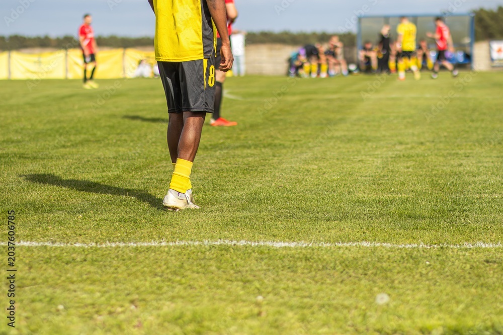 Obraz premium Male soccer player in yellow jersey and black shorts stands on green grass field, teammates and opposing players visible in the background during a match