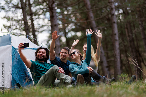 Friends enjoying a camping trip in the forest while taking a selfie together with smiles and waves