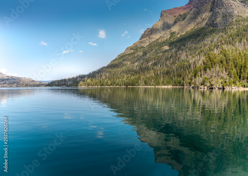 Turquoise Saint Mary Lake surrounded by steep forested mountain slopes and weathered tree stumps under a bright blue Montana sky in Glacier National Park