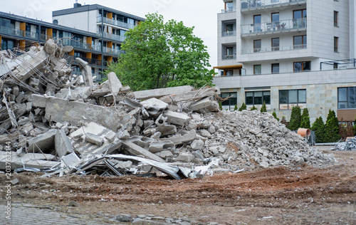 Photography A large pile of broken concrete rubble and twisted metal rebar from a demolished structure sitting in front of a modern residential apartment building
