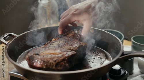 Hand lifts a sizzling, seared steak from a hot pan. Smoke rises as the meat cooks in oil. Close-up of juicy beef being flipped. Ideal for food, cooking, or restaurant marketing videos