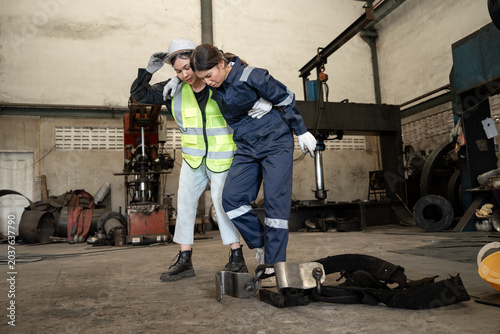 Young Asia female engineer with hurting knee sitting on the floor and female worker in safety helmet helping and calling ambulance