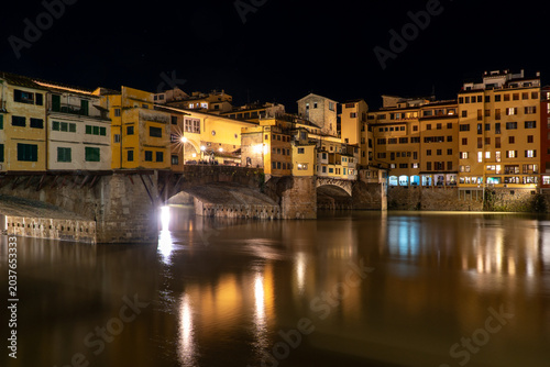 The historic Ponte Vecchio bridge in Florence, Italy, illuminated at night with reflections shimmering on the Arno River