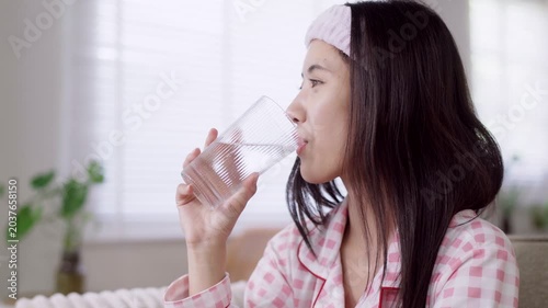 young Asian women morning routine drinking water from glass wearing pink checkered pajamas headband refresh hydration health wellness lifestyle at bright home