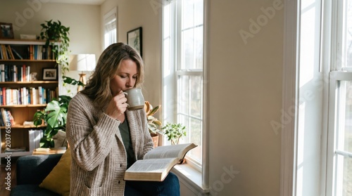 Middle-aged woman drinking coffee while holding open Bible by window in cozy home library