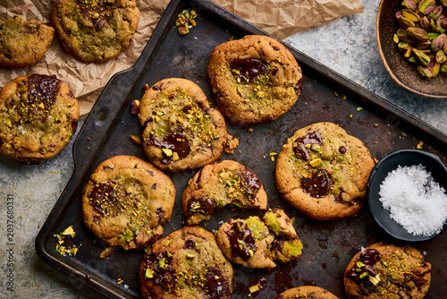 Pistachio-studded chocolate cookies with molten centers arranged on a dark, weathered baking tray, with scattered nuts, flaky salt, parchment paper, and a ceramic bowl of pistachios.