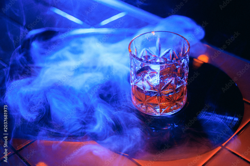 Obraz premium Blue smoke whiskey glass on vinyl record with dark neon nightlife mood. Concept of smoke drink hero shot, premium whiskey campaign, music bar poster, speakeasy mood, and social cover.