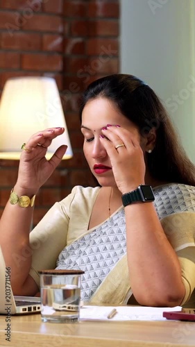 Drinking water while suffering from stress and headache, Indian young businesswoman in ethnic saree sitting at cozy office desk with laptop trying to stay hydrated during long working hours