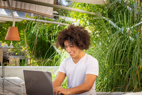Smiling young ethnic men , dressed in casual clothes, works on a laptop outdoors, in a green area in nature. Green open space for coworking, freelancer, relaxing, coffee, food. Free place to work.