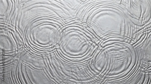 Water ripples create concentric circular patterns across the smooth surface of a dark liquid pool where gentle disturbances cause shifting geometric reflections and rhythmic fluid motion throughout.