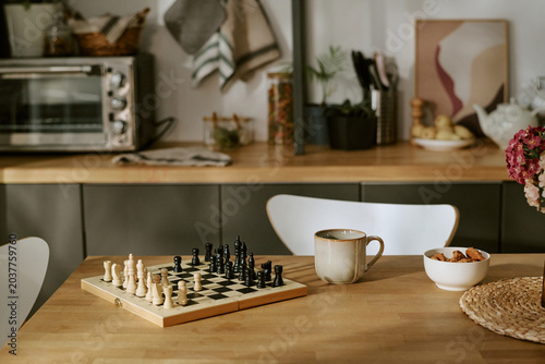 Wide shot of chessboard standing on kitchen table beside coffee mug and snack bowl, suggesting strategic thinking and leisure. Useful for lifestyle, home routine, planning, or hobby marketing