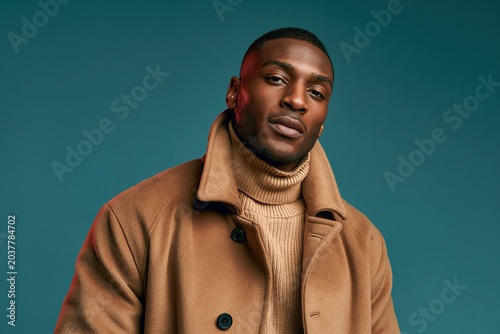 Stylish Black man wearing a camel coat and turtleneck sweater against a teal background