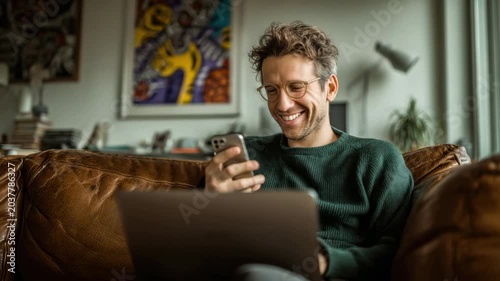 Man smiles while using smartphone and laptop in cozy living room with art on wall