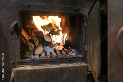 Close up of burning wooden logs blazing in fire flames inside wood burning stove with open glass door in cosy warm home living room.
