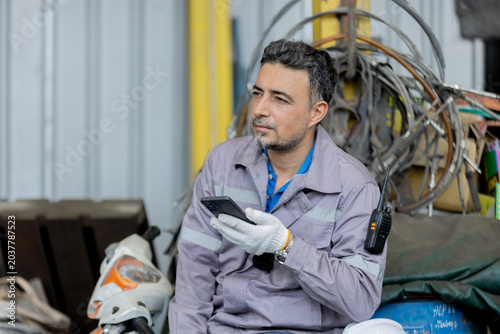 Bilde på lerret A worker in a grey uniform and gloves sits in an industrial setting, holding a smartphone and looking thoughtfully to the side