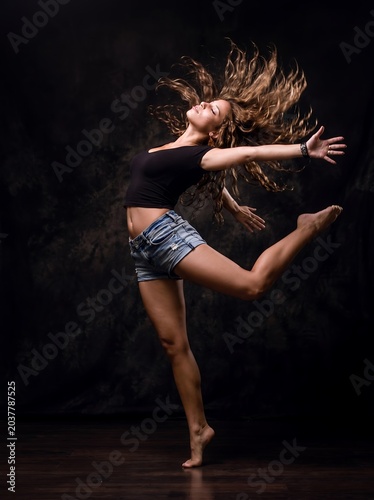 Dynamic action shot of a beautiful young female dancer leaping mid-air with flying long curly hair in front of a dark dramatic textured studio background.