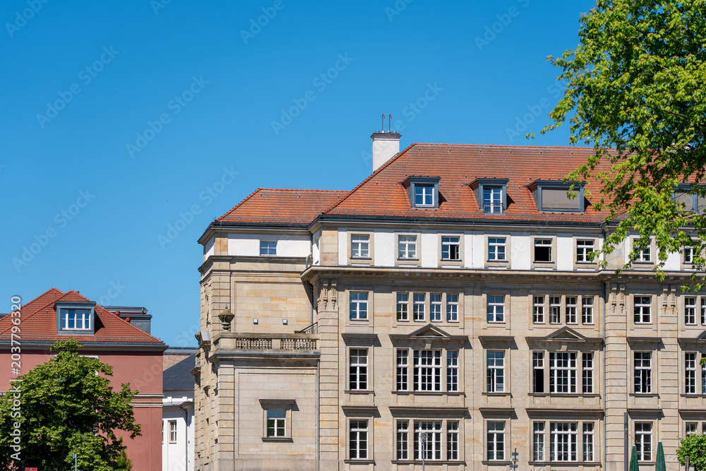 Fototapeta premium Historic stone facade with red tile roof stands under a clear blue sky. Green deciduous tree foliage frames the urban architecture.