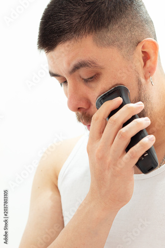 Portrait of a young man running a portable electric shaver over his cheeks to trim his beard. Concept of personal grooming