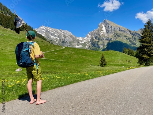 Barefoot boy hiking through rural Toggenburg, Switzerland, carrying a wooden stick over his shoulders with hiking shoes dangling from it.