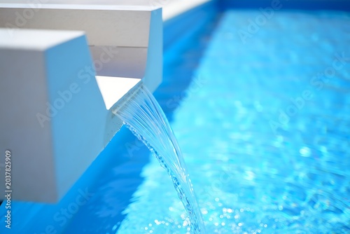 Closeup of elegant stone fountain streaming clean water into luxury swimming pool under bright sunlight, creating refreshing ripples, tranquil ambiance, and modern outdoor relaxation atmosphere.