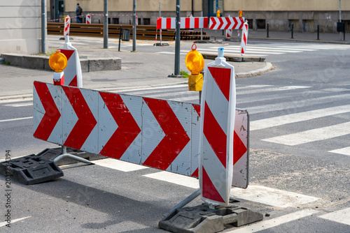 Road barriers with white and red stripes and warning signs with chevrons and yellow lamps, placed on asphalt near a pedestrian crossing during public construction works in the city, photographed in da