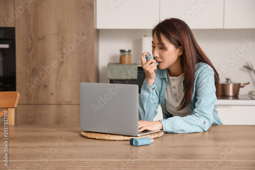 Tableau sur toile Young Asian woman with laptop using inhaler at table in kitchen