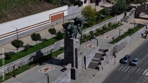 An aerial shot of the historic equestrian monument situated next to a busy city road with cars and a river on a bright sunny day in Kapan, Armenia.

