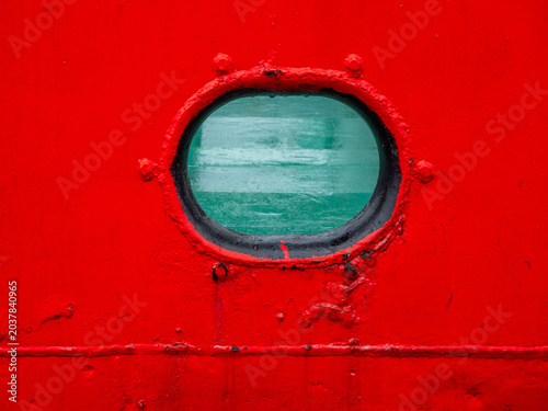 Oval ship porthole window on bright red metal hull background.  Close-up of a classic nautical porthole on a vessel side painted in vibrant red. Through the glass, green deck or sea surface is visible
