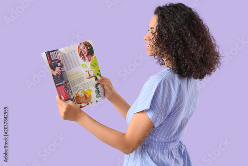 Young African-American woman reading fashion magazine on lilac background