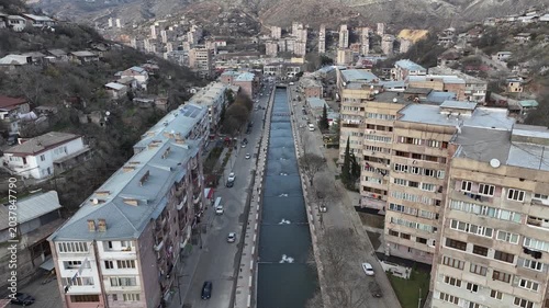 An aerial drone shot of a beautiful, scenic city nestled in a deep mountain valley with residential buildings, houses, and roads on a sunny day in Kapan, Armenia.

