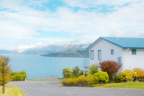 Konstfotografi South Island, New Zealand: charming white holiday home with blue roof, vacation house with calm turquoise sea bay and distant volcanic mountain view
