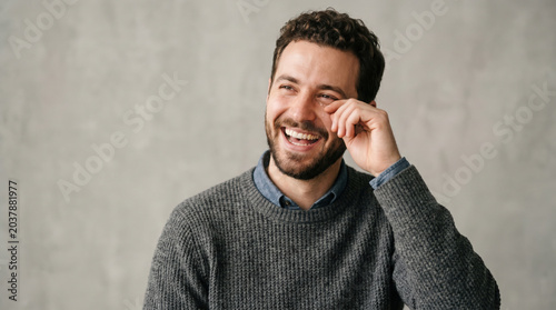 Bearded man with curly hair laughs joyfully, wiping a tear from
