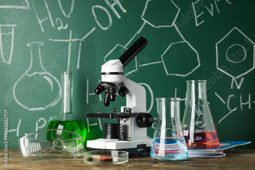 Modern microscope, petri dish and flasks on wooden table near chalkboard with...
